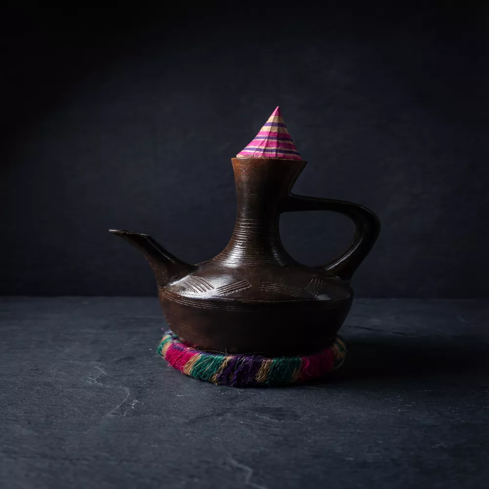 دلة قهوة سودانية (جبنة) مصنوعة من الفخار البني المصقول مع قاعدة منسوجة يدوياً بألوان زاهية وتصميم تقليدي أنيق